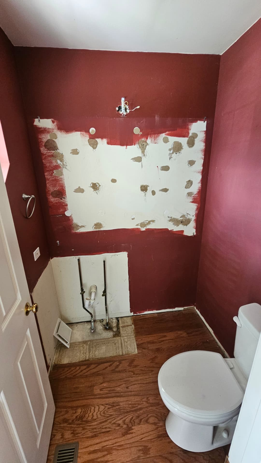 Powder room before work with dark red walls, patched drywall and exposed plumbing